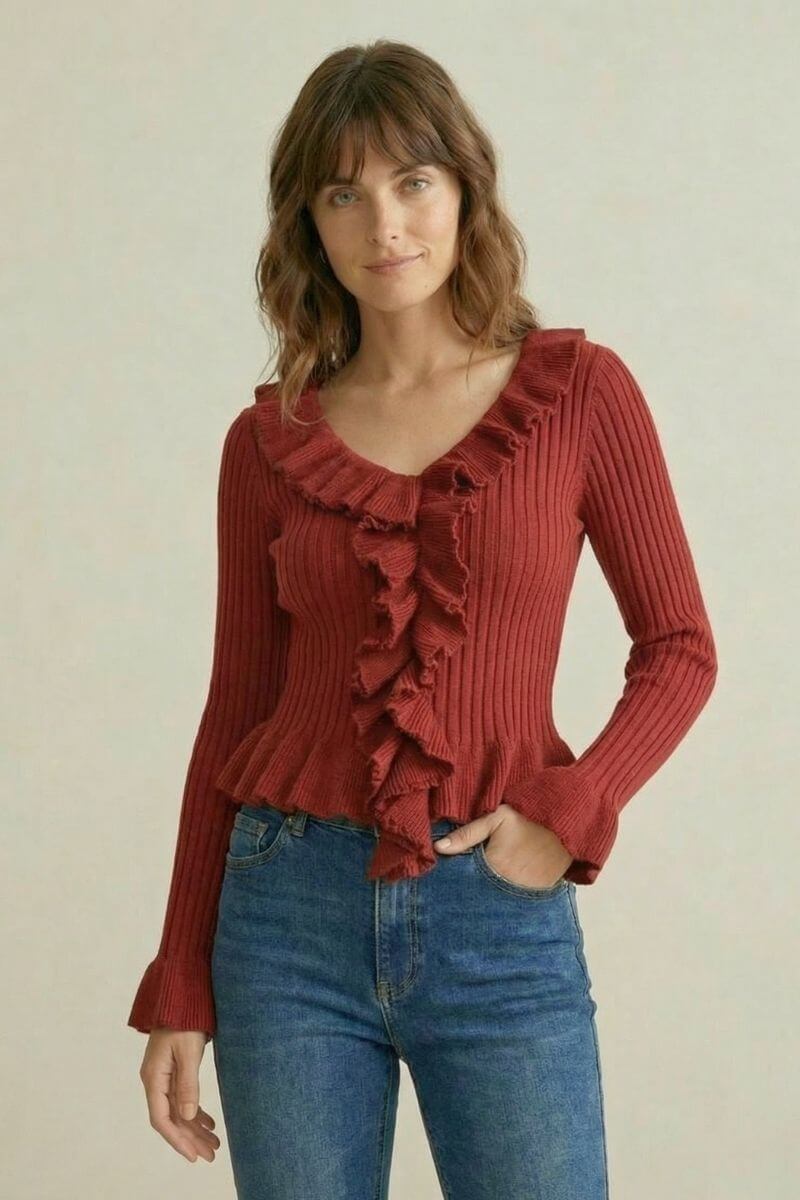 Woman wearing a red ribbed knit top with cascading front ruffles and long sleeves paired with blue jeans, neutral background.