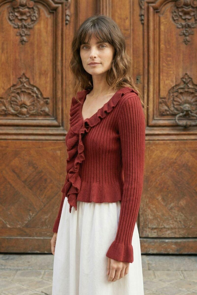 Woman standing outdoors in front of carved wooden doors wearing a red ribbed knit ruffle top and white skirt.