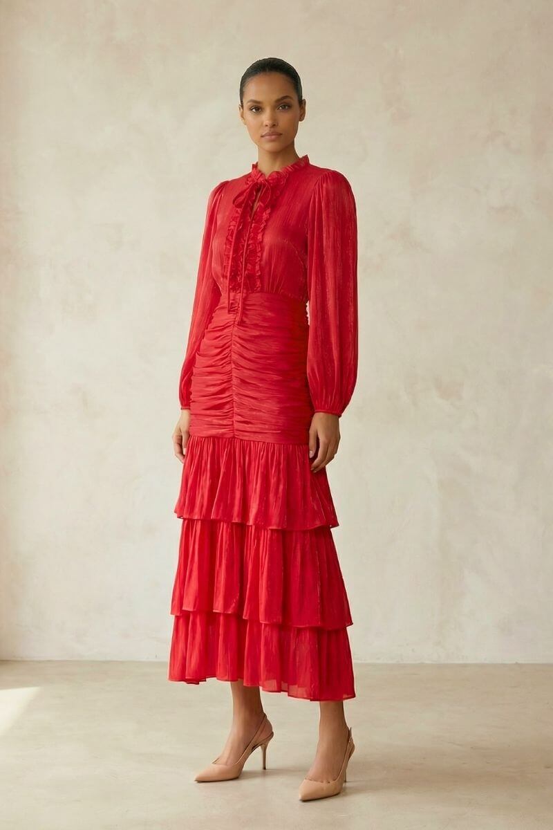 A model stands against a soft neutral backdrop wearing a red long-sleeve midi dress with a ruffled high neckline, gathered waist, and layered tiered skirt, styled with nude pointed-toe heels for an elegant, romantic look.