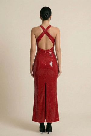Back view of a red sequin halter maxi dress featuring crisscross straps, an open back, and a center back slit, worn by a model in black heels on a neutral studio set.