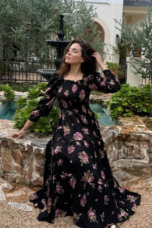 Model posing outdoors beside a fountain in a black smocked floral maxi dress with voluminous sleeves and rose pattern, hand brushing her hair as she faces the breeze in a tranquil garden atmosphere.