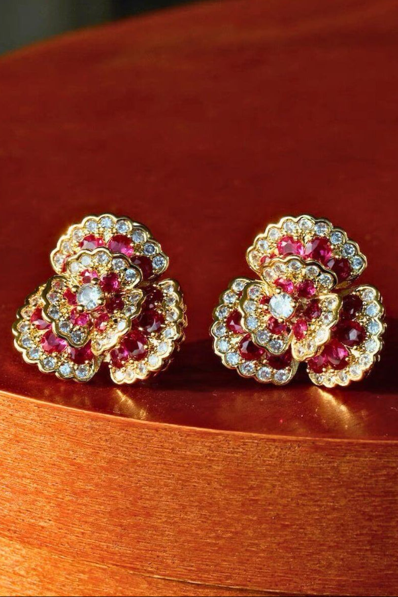 A pair of ruby and clear crystal flower stud earrings in gold setting, photographed symmetrically against a black background.