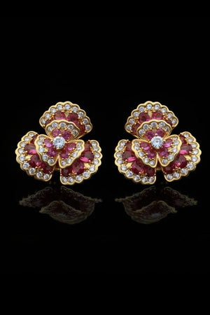 A close-up of ruby and clear crystal floral cluster stud earrings set in gold, displayed on a warm wooden surface with rich red tones.