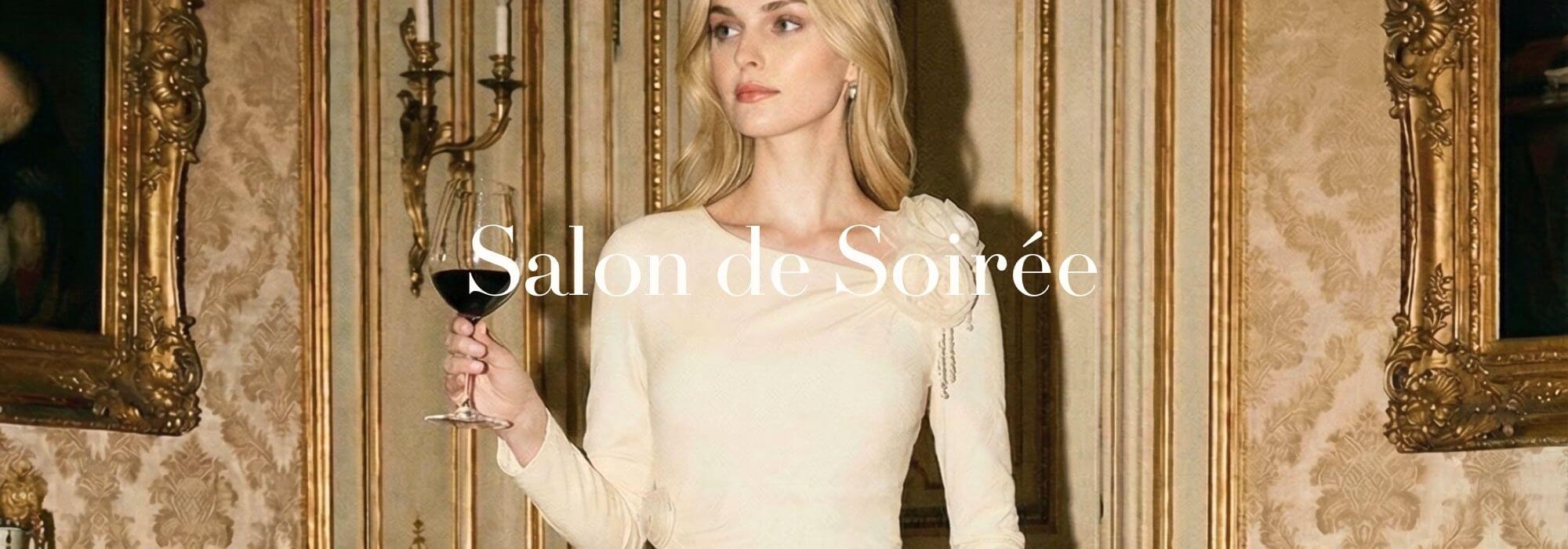 A blonde female model stands in a gilded Parisian salon holding a glass of red wine, wearing an ivory one-shoulder fitted evening dress with a soft floral appliqué, evoking refined candlelit elegance.