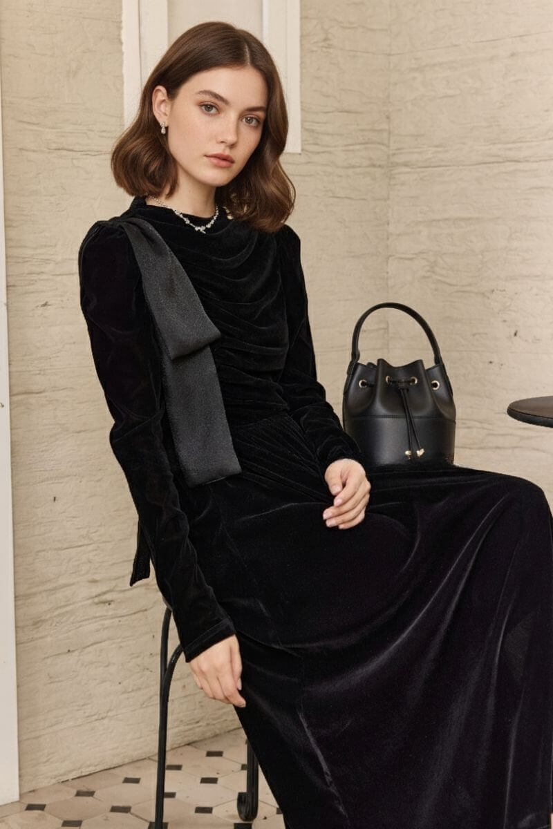Model seated by a small café table wearing a black velvet maxi dress with a softly ruched bodice and shoulder bow, accessorized with a pearl necklace; black bucket bag rests in the background.