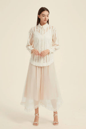Model wearing a sheer ivory lace blouse with long sleeves and a collared neckline, styled with a flowing tulle midi skirt.