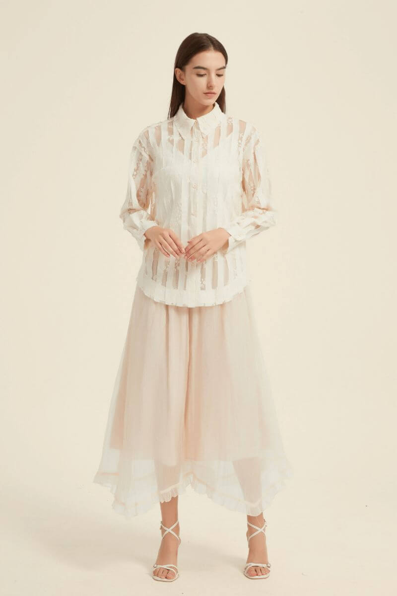 Model wearing a sheer ivory lace blouse with long sleeves and a collared neckline, styled with a flowing tulle midi skirt.