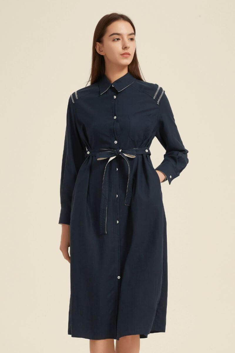 Side angle of a navy collared midi shirt dress with front buttons and waist tie, styled with brown ankle boots for a polished modern look.