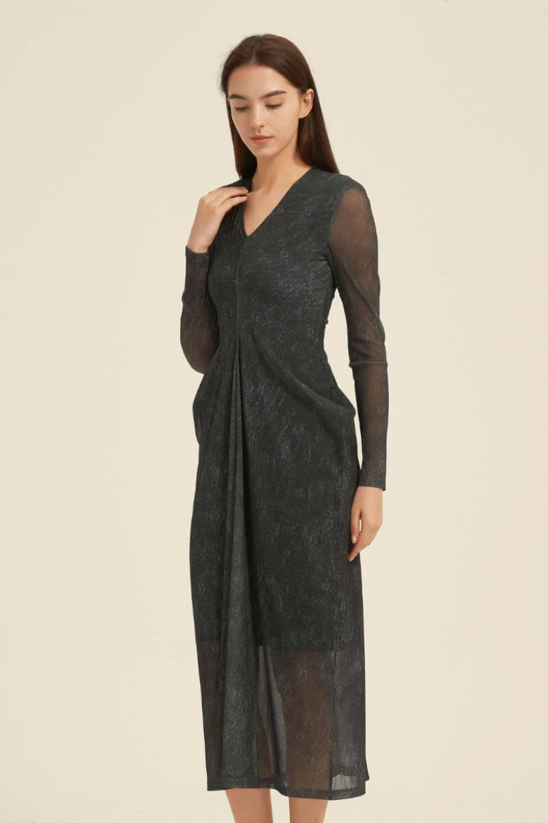 Angled view of a black midi dress showing the V-neck bodice, sheer long sleeves, draped front pleat, and calf-length semi-sheer skirt.