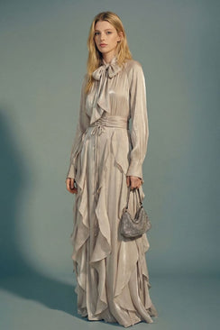 A model styles a silver satin ruffle maxi dress with a bow neckline, long sleeves, and defined waist, paired with a small handbag for an elegant, refined look.