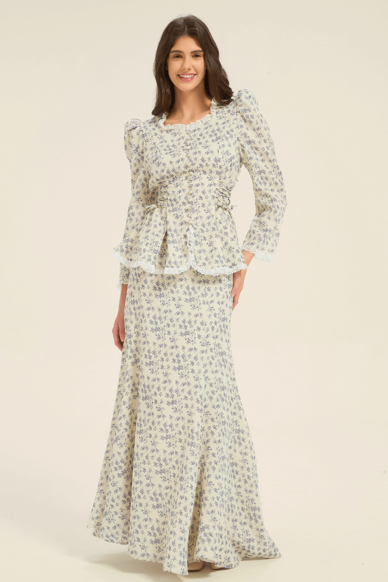 Model smiling while wearing a cream and blue floral peplum blouse with puff sleeves and matching maxi skirt, front view.