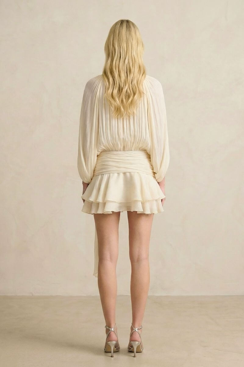 A model wearing a soft pearl pleated chiffon mini dress set photographed at an angle, emphasizing the fluid sleeves, wrap waist tie, and lightweight chiffon movement.