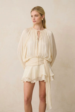 A model stands facing forward in a soft pearl pleated chiffon mini dress set, featuring a tie-neck blouse, softly gathered waist, and flowing ruffle mini skirt.