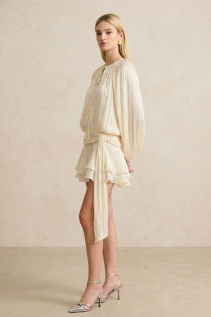 A model wearing a soft pearl pleated chiffon mini dress set shown from the side, highlighting the airy drape, balloon sleeves, wrap waist tie, and layered ruffle hem.