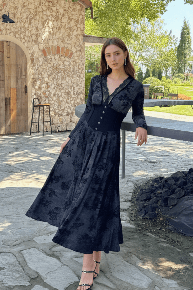 Model in a flowing black velvet dress with a corset waist, long sleeves, and deep neckline, posing gracefully outdoors against a stone wall and garden backdrop.