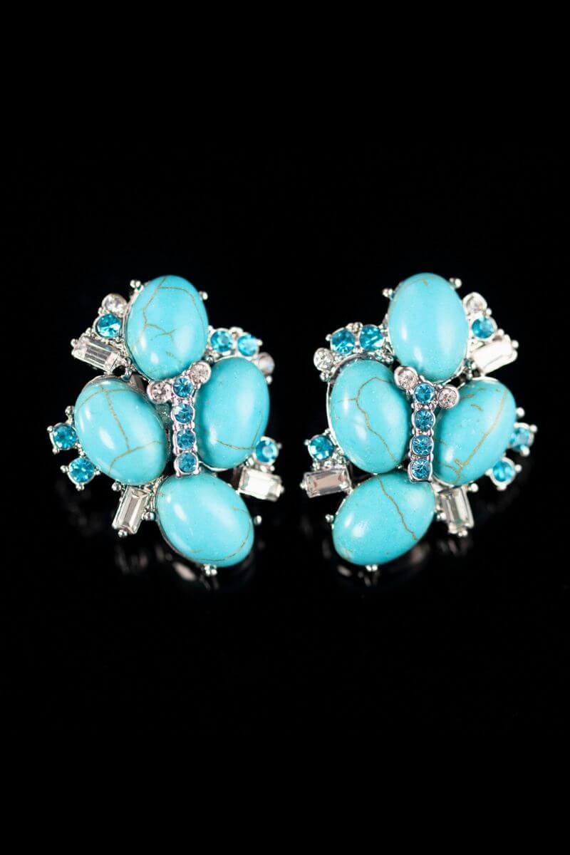 A pair of turquoise cluster stud earrings displayed on a black reflective surface, showcasing the symmetrical floral design with turquoise stones and crystal details.