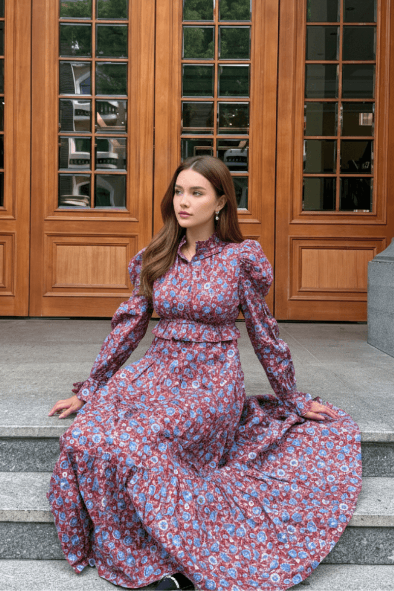 Model sitting on stone steps wearing a red and blue floral print maxi dress with puff sleeves and a high ruffled neckline.