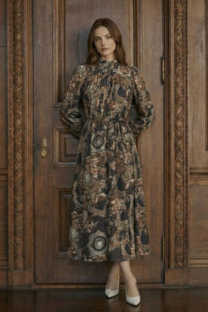 A woman stands full-length in a vintage-inspired floral midi dress with long sleeves, a cinched waist, and flowing skirt, styled with neutral heels in front of carved wood doors.