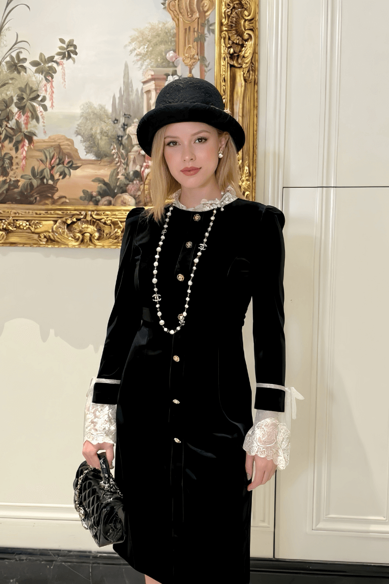 Elegant woman in a black velvet midi dress with gold buttons, lace cuffs, and pearl necklace, styled with a black hat indoors.