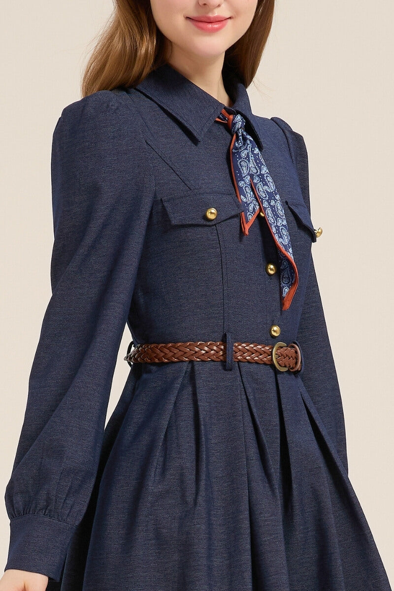 Close-up of a navy twill midi dress featuring gold button details, chest pockets, a woven brown belt, and a blue paisley scarf tied at the collar.