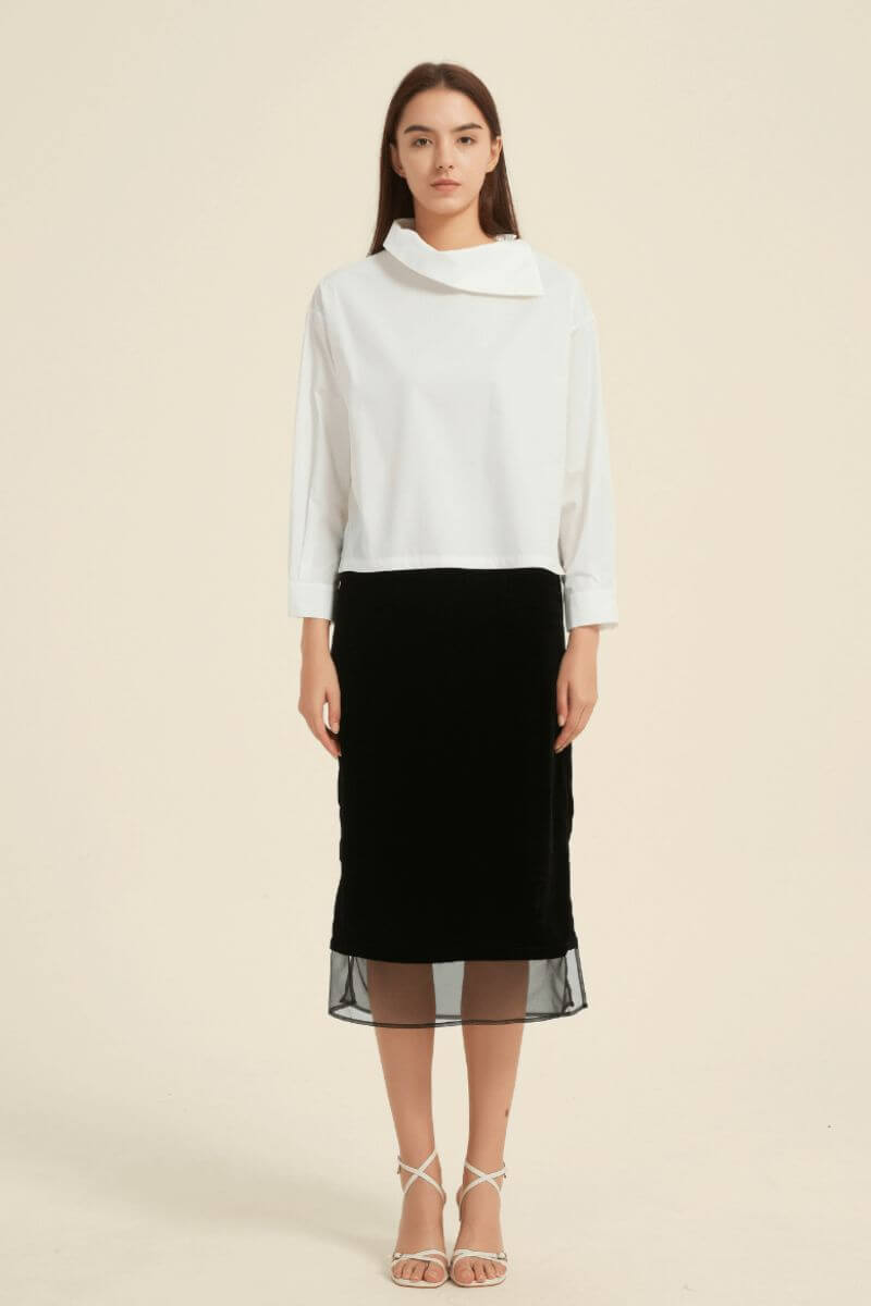 Model wearing a white asymmetric-collar blouse tucked into a black midi skirt with sheer layered hem — elegant contrast look from Sioéne.