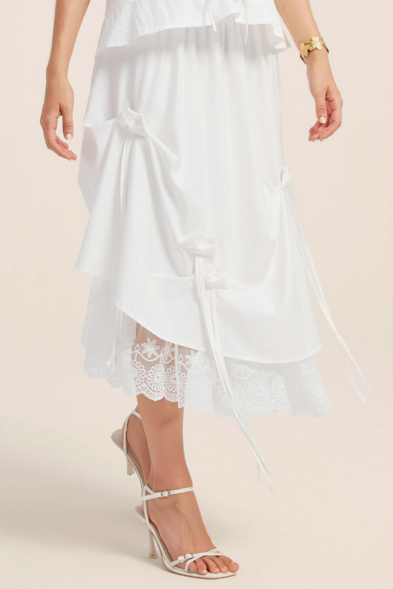 Close-up of a white midi skirt showcasing cascading satin bows, asymmetrical draping, and delicate lace trim at the hem.