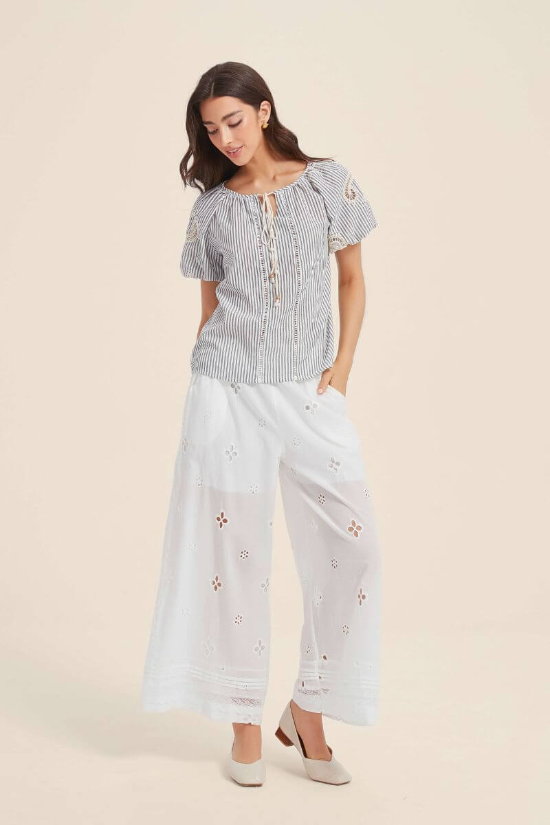 Woman wearing white embroidered wide-leg pants with floral cutouts and a navy striped lace-trim top, styled with beige heels for a light summer look.