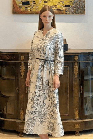 Lifestyle image of a woman in a white floral-print midi dress styled with a slim black belt and three-quarter sleeves in an elegant interior.