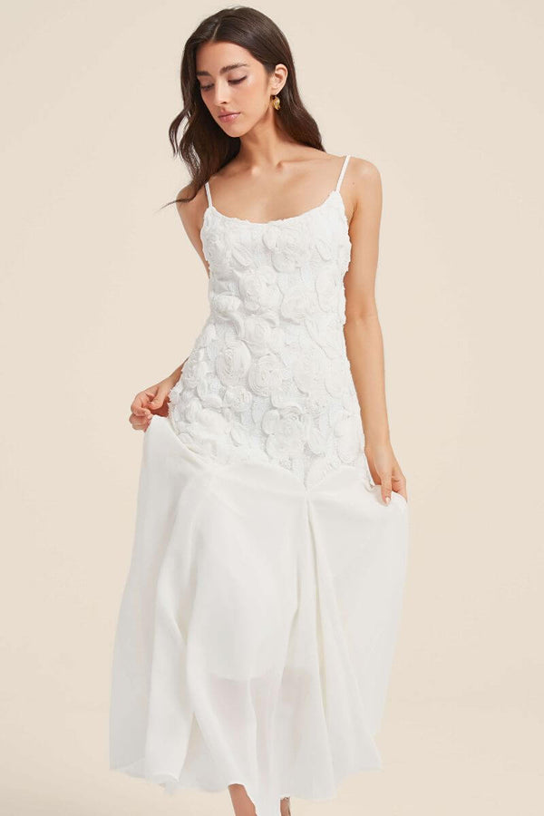 Woman wearing a white floral appliqué maxi dress with slim straps and rosette lace details, gently lifting the skirt to reveal its airy chiffon layers.