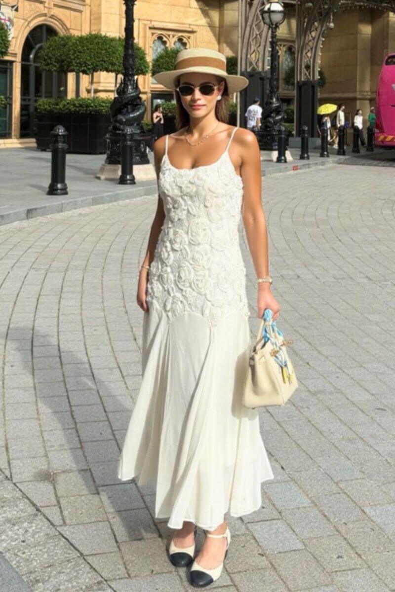 Woman wearing a white floral lace maxi dress with spaghetti straps styled for summer street fashion, paired with a straw hat, beige bag, and two-tone espadrille heels.