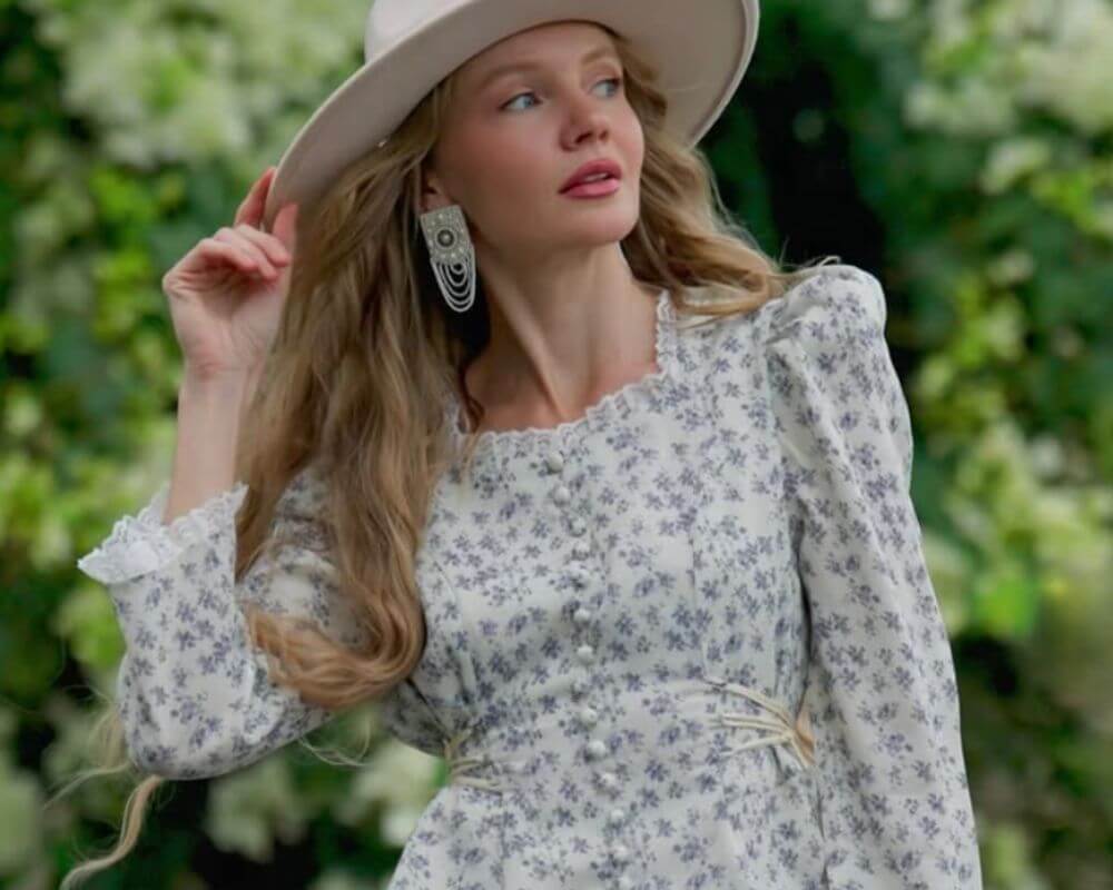 A woman wearing a white puff-sleeve maxi dress with blue floral print and front button details, paired with a wide-brim hat and statement earrings, in a garden setting. A romantic cottagecore-style look by Sioéne.