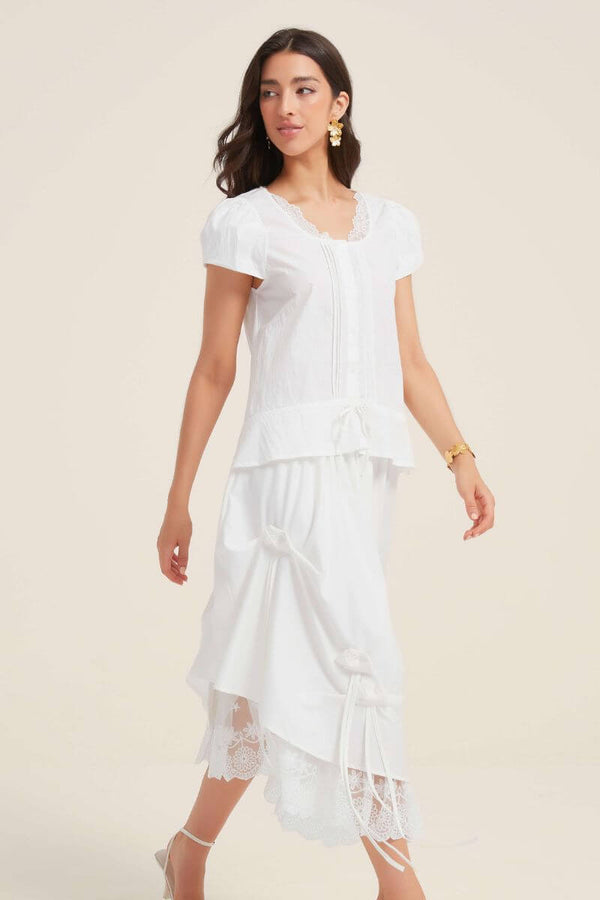 Woman wearing a white lace-trim blouse with short sleeves paired with a draped midi skirt featuring satin bows and lace underlay, creating a romantic monochrome look.