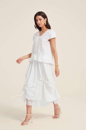 Side view of a woman wearing a white lace-trim blouse and matching bow-detail skirt with layered lace hem, styled with gold floral earrings and white heels.