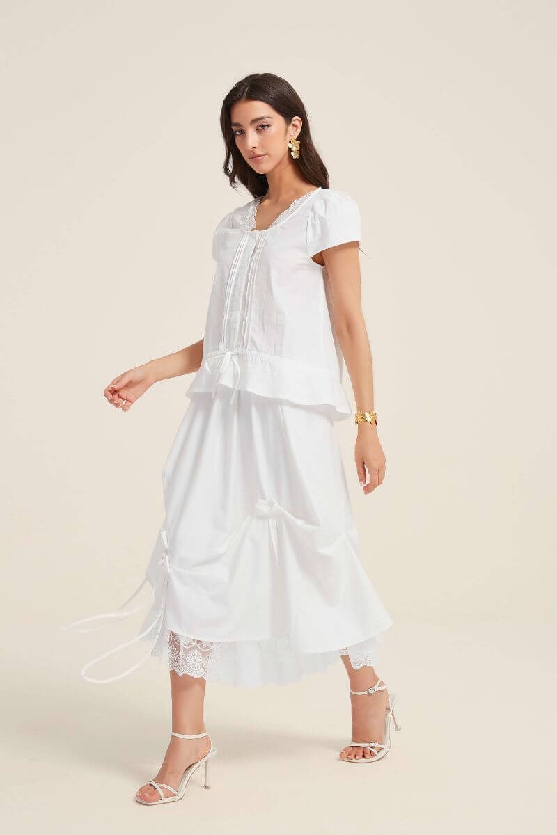 Side view of a woman wearing a white lace-trim blouse and matching bow-detail skirt with layered lace hem, styled with gold floral earrings and white heels.