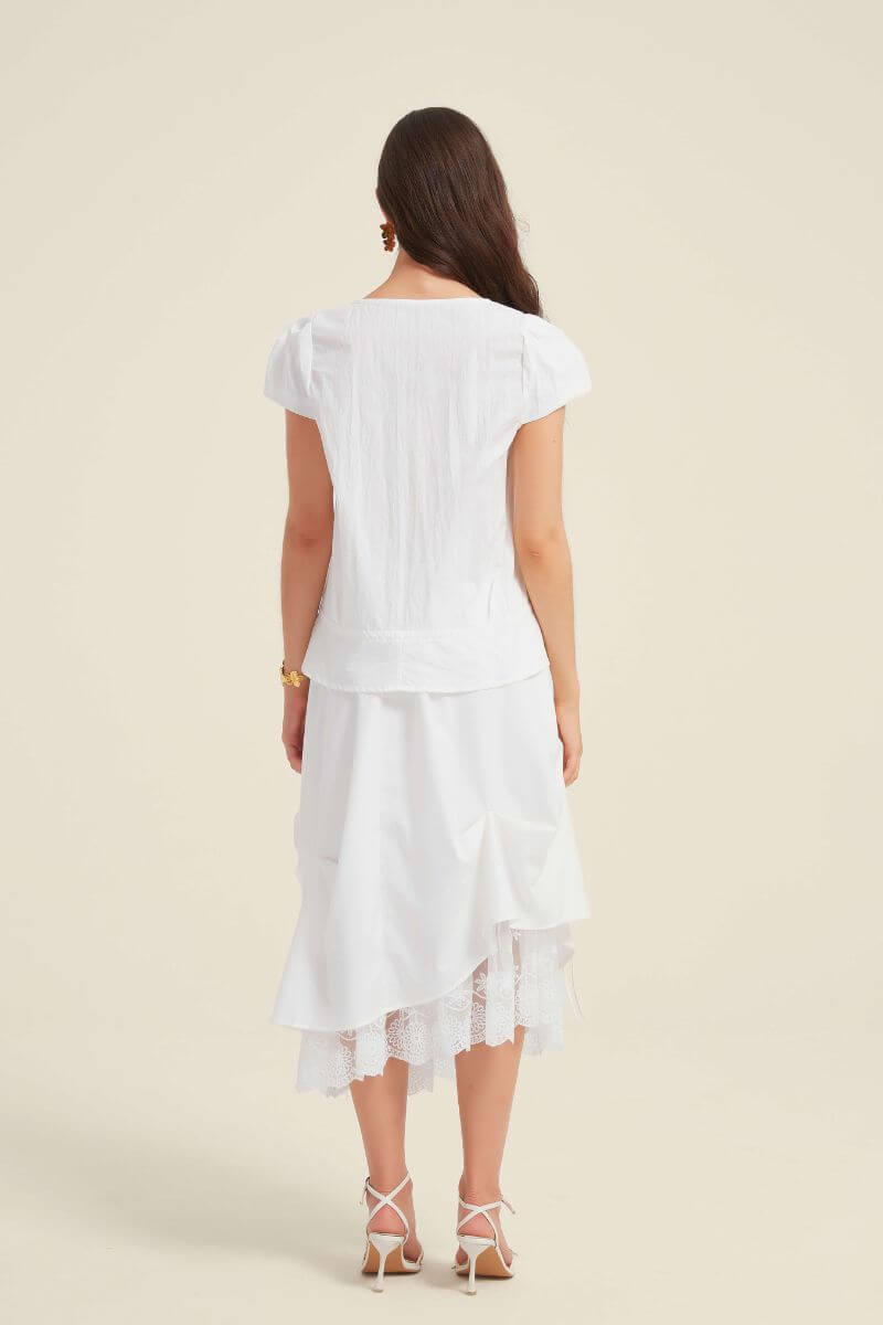 Back view of a woman wearing a white lace-trim blouse with puff sleeves and a peplum hem, styled with a white layered lace midi skirt.