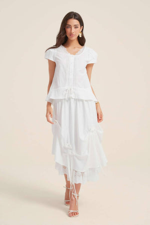 Full-length view of a woman in a white lace-trim drawstring blouse paired with a tiered white skirt with satin bows and lace hem, evoking a vintage romantic mood.