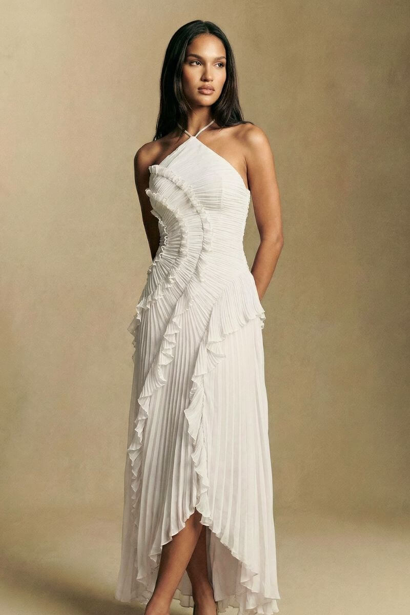 Model wears a white pleated halter dress with an asymmetrical hemline, the layered ruffles moving gently as she stands against a neutral backdrop.
