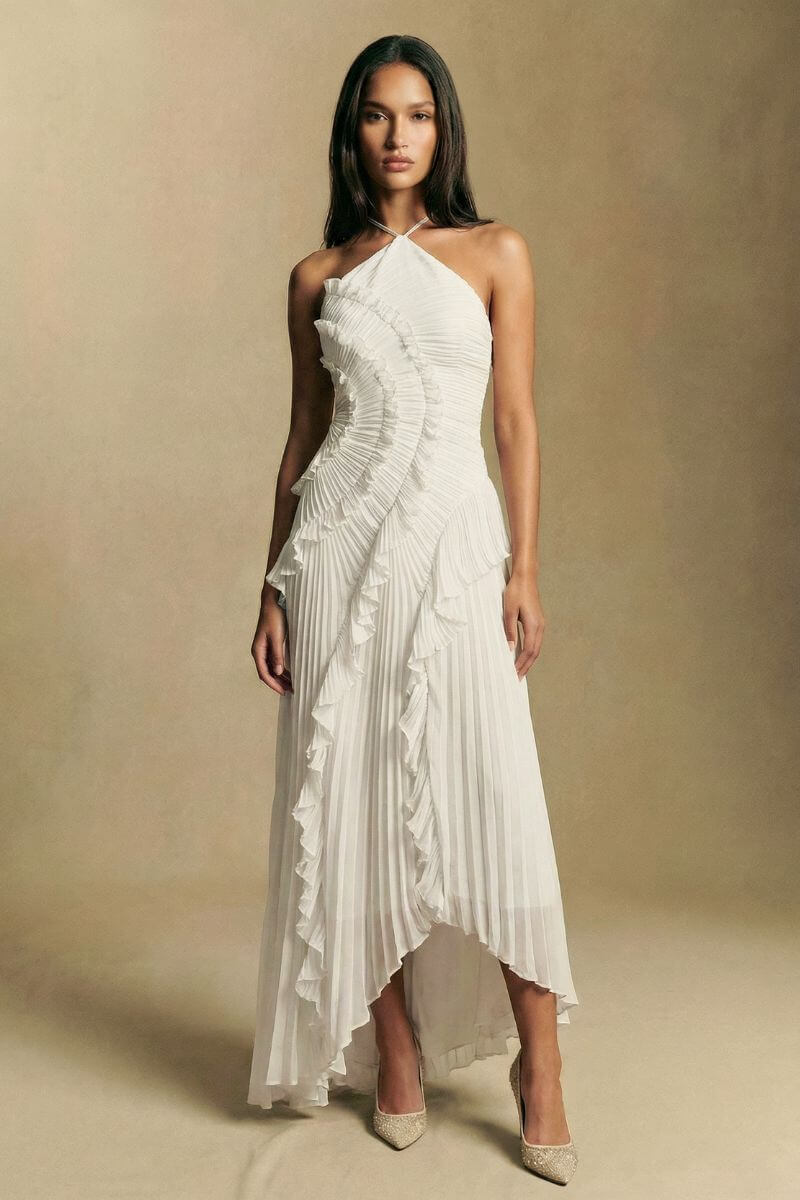 Model stands facing forward in a white pleated halter midi dress with soft layered ruffles, styled with pointed heels against a warm neutral studio backdrop.