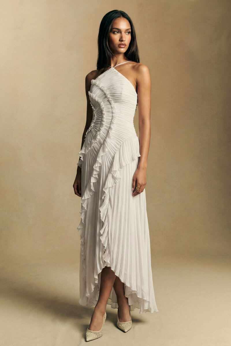 Model poses in profile wearing a white pleated halter midi dress with flowing ruffles that create a soft, sculpted side silhouette.