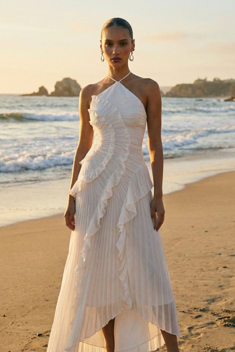 Model wears a white pleated halter midi dress with cascading ruffle details, standing on a sandy beach at sunset with ocean waves behind her.