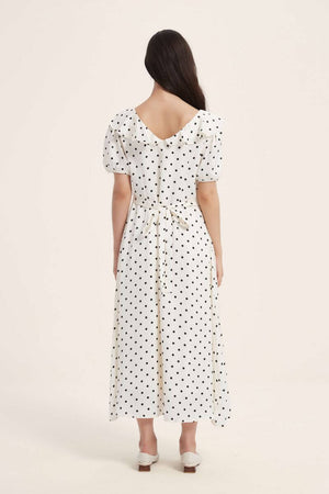 Woman wearing a white polka dot midi dress with puff sleeves, ruffled neckline, and a bow tie at the waist, shown from the back against a neutral background.