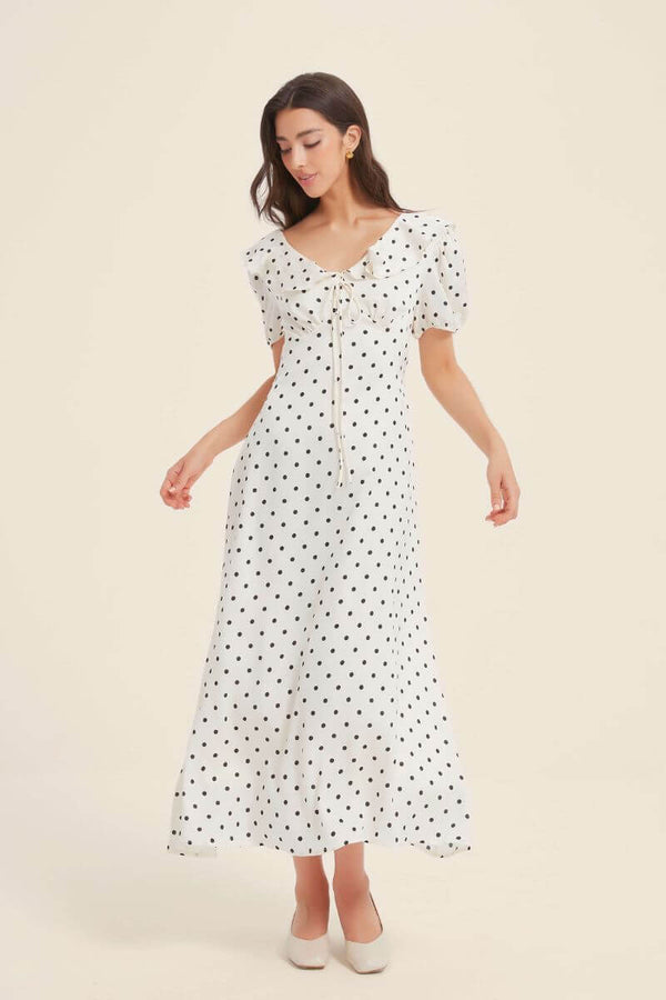 Woman modeling a white midi dress with black polka dots, puff sleeves, and a ruffled drawstring neckline, styled with cream block heels.