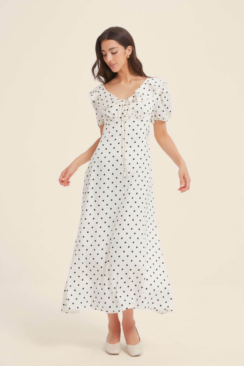 Woman modeling a white midi dress with black polka dots, puff sleeves, and a ruffled drawstring neckline, styled with cream block heels.