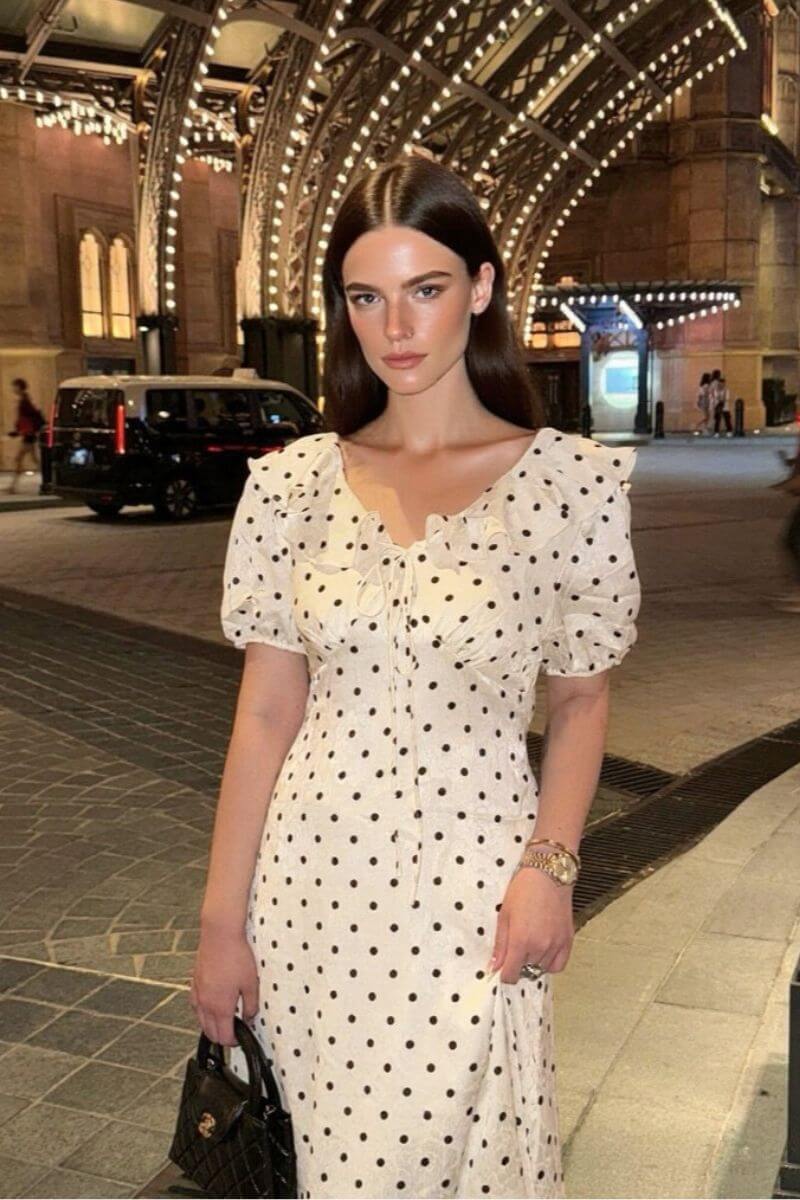 Woman wearing a white puff sleeve polka dot midi dress at night under city lights, styled with a black handbag and gold watch for a chic Parisian look.