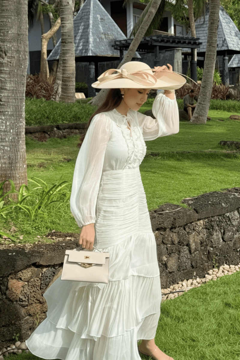 Woman in a flowing white maxi dress with ruched detailing, paired with a dramatic wide-brimmed sun hat, walking in a tropical outdoor setting.