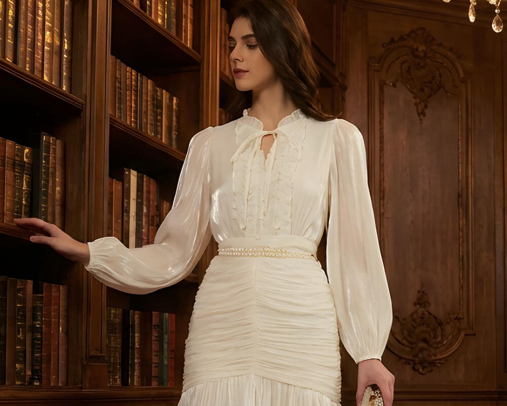 Woman wearing a white ruffle long-sleeve maxi dress with a ruched waist and pearl belt, standing in a vintage wooden library and touching a bookshelf.
