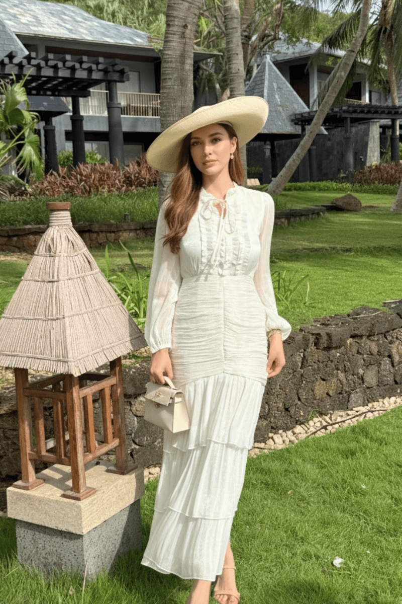 Woman in a long-sleeve white tiered dress with delicate ruffle details, carrying a cream handbag and wearing a wide sun hat in a garden resort.