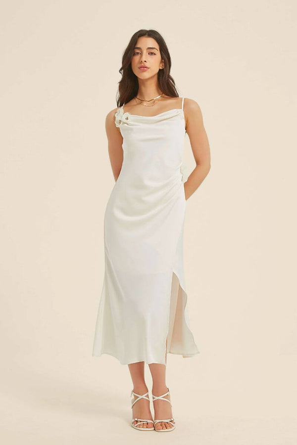 Woman wearing a white satin midi dress with a draped cowl neckline and floral appliqué on the shoulder and hip, styled with white heels.