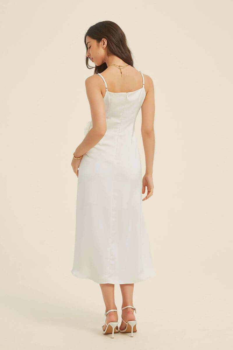 Back view of a woman in a white satin midi dress with thin straps and a smooth bias-cut silhouette, styled with gold jewelry and heels.