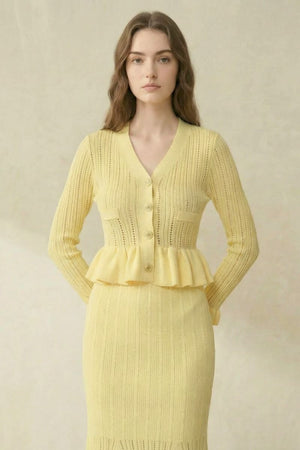 Close-up of a yellow knit peplum cardigan with vertical pointelle knitting, covered buttons, and ruffled hem in a neutral studio.